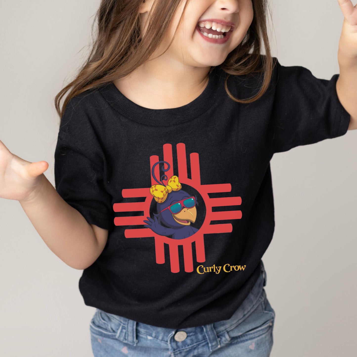 Smiling school-age child wearing a bright Curly Crow Zia t-shirt, proudly showing off the colorful crow design inspired by the Zia sun symbol.