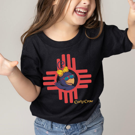 Smiling school-age child wearing a bright Curly Crow Zia t-shirt, proudly showing off the colorful crow design inspired by the Zia sun symbol.