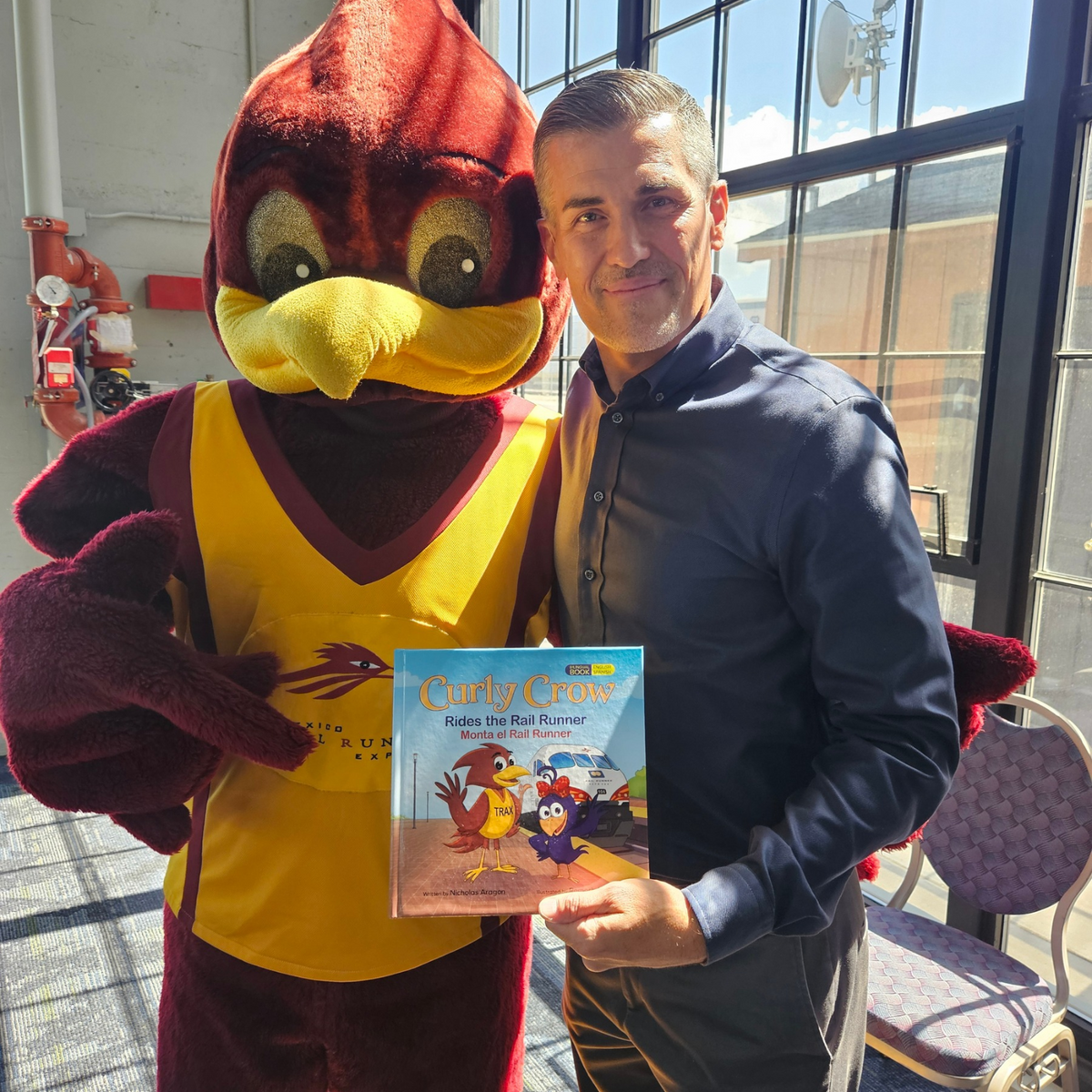 Author Nicholas Aragon standing next to the Rail Runner mascot Trax while holding the popular kids book Curly Crow Rides the Rail Runner
