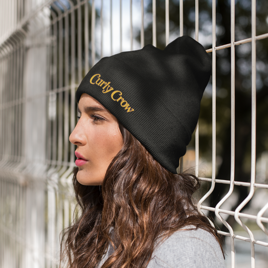 Woman wearing a black beanie with 'Curly Crow' text, standing against a wire fence.